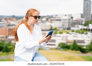 A young woman enjoys a sunny day in the city while using her smartphone on a scenic overlook - Powered by Shutterstock