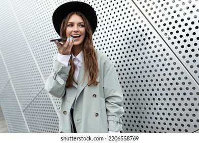 A Young Woman Entrepreneur In An Autumn Coat And A Black Hat Speaks On A Mobile Phone With A Speakerphone Against The Background Of An Aluminum Facade Of A Business Center