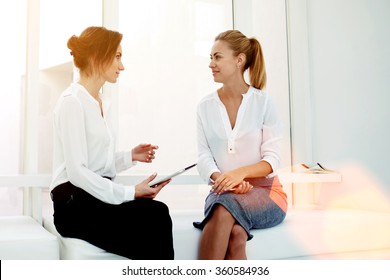 Young woman entrepreneur hold touch pad while telling something to her partner during business meeting, two female confident proud CEO discussing new project while sitting in modern office interior 