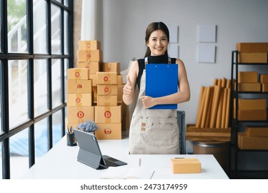 Young woman entrepreneur in home office with shipping boxes, holding clipboard and giving thumbs up, representing small business and e-commerce. - Powered by Shutterstock