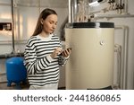 A young woman examines a non-operational boiler with concern, holding a phone in her hand, likely seeking help or planning to call for repair services. The basement setting indicates a residential