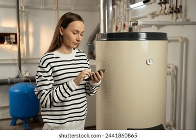 A young woman examines a non-operational boiler with concern, holding a phone in her hand, likely seeking help or planning to call for repair services. The basement setting indicates a residential