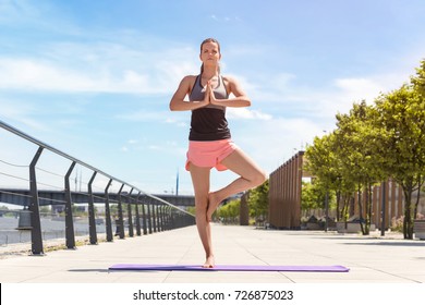 Young Woman Exercise Yoga In The City Hands Up And Standing On One Leg In Sunny Day. 