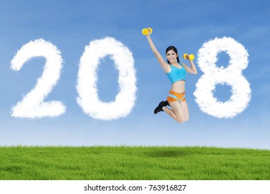 Young woman exercising with two dumbbells in the park while leaping with clouds shaped numbers 2018 in the sky