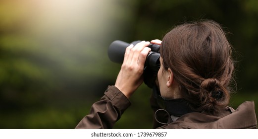 Young woman explorer with binoculars and telescope watching for animal in the wild forest. Blur dark background. The concept of hunting, travel and outdoor recreation.