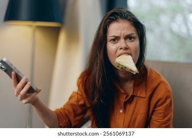 Young woman expressing frustration while holding a smartphone and biting a sandwich, showcasing a relatable moment of multitasking in a modern lifestyle - Powered by Shutterstock