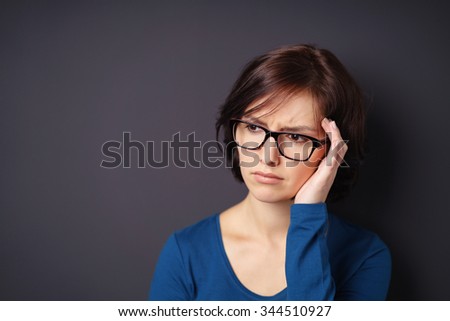 Young Woman with Eyeglasses Showing Stressed Face While Looking Away Against Gray Wall with Copy Space on the Left.