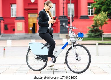 Young woman in eyeglasses uses smartphone while riding a bicycle in the city. Woman in smart casual wear uses bike for getting at work
