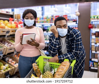 Young Woman In Face Mask Checking Grocery List At Supermarket, Her Boyfriend Sick And Tired Of Shopping. Millennial Couple Purchasing Foodstuff At Modern Mall