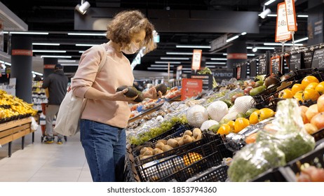 Young Woman In Face Mask Choosing Avocado At Fresh Fruit And Vegetable Section In Supermarket. Preventive Measures In Grocery Shop To Limit Covid 19 Spread. Warsaw-Poland-2020