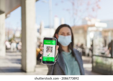 Young Woman With Face Mask Showing The Covid Passport In Her Cell Phone. Selective Focus.