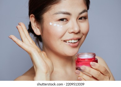 Young Woman With Facial Cream Jar And Eye Cream On Face Isolated On Blue Background.