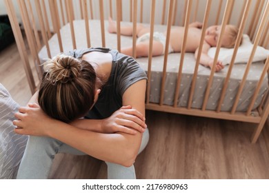 Young woman facing difficulties of maternity sitting next to baby crib with head on her knees crying with exhaustion and desperation, feeling lonely. Postnatal depression symptoms