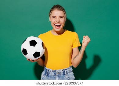 Young woman fan wear basic yellow t-shirt cheer up support football sport team hold in hand soccer ball watch tv live stream do winner gesture celebrate clench fist isolated on dark green background - Powered by Shutterstock