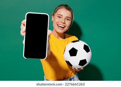 Young woman fan wear basic yellow t-shirt cheer up support football sport team hold soccer ball watch tv live stream use mobile cell phone closeup blank screen isolated on dark green background studio - Powered by Shutterstock