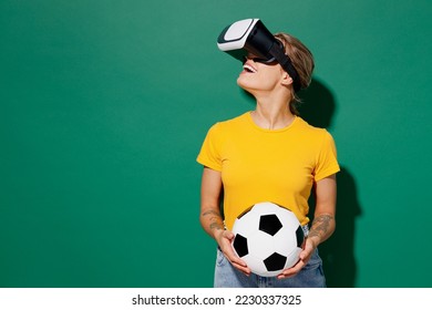 Young woman fan wears yellow t-shirt cheer up support football sport team hold soccer ball watch tv live stream watching in vr headset pc gadget look aside on area isolated on dark green background - Powered by Shutterstock