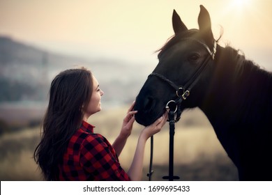 Young Woman Farmer In A Red Checkered Shirt Hugs Her Black Horse At Sunset - Concept Of Love Between People And Animals