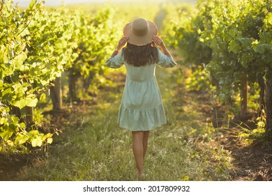 A young woman with fashion accessories stands in a vineyard field in a green summer dress and holds a straw hat with her hands in sunny day, view from the back. Summer inspiration.