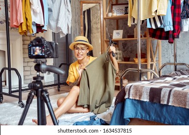 Young woman fashion blogger influencer wearing hat and eyeglasses sitting on fluffy carpet at urban stylish apartment recording video vlog on digital camera telling clothing tips positive