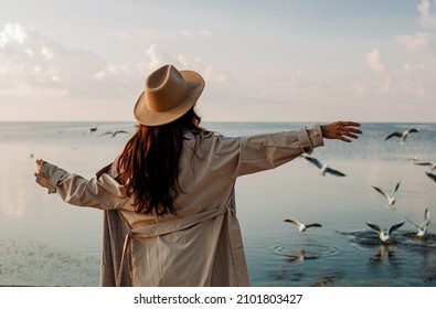 Young woman feeds seagulls at winter sea beach. Amazing coastline scene with girl. Concept of freedom, travel, flying. Lifestyle moment.