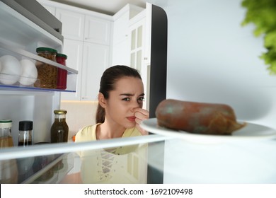 Young Woman Feeling Bad Smell Of Spoiled Sausage In Refrigerator, View From Inside