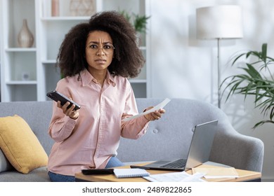 Young woman feeling stressed while sitting on couch holding phone and papers. Worried expression suggests financial or work-related issues. Home office setup with laptop and documents on coffee table - Powered by Shutterstock