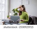 Young woman feeling unwell and measuring her temperature with a thermometer while sitting on a sofa at home