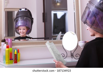 Young Woman Female Client Reading Magazine And Drinking Hot Beverage Coffee Tea In Hairdressing Beauty Salon. Girl In Hair Rollers Curlers With Hairdryer Dryer Relaxing By Hairdresser Hairstylist.