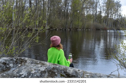 Young woman female in green sweater and hat sit on rock resting, looking at the lake while drinking water from metal bottle. Mindfulness in nature. Cold weather lake shore. - Powered by Shutterstock