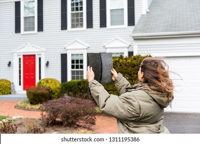 Young Woman Female Homeowner Standing In Front Of House On Windy Day In Coat Jacket During Winter Storm Holding Up Roof Tile Shingle Inspecting Damage