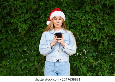 Young woman in festive outfit holding smartphone against green background during holiday season. Caucasian woman in Santa hat with phone in hands on green background. Holiday is coming to us. Gifts. - Powered by Shutterstock