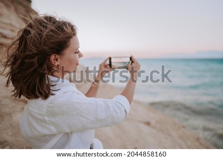 Young woman filming video with mobile phone on beach near blue sea. Girl using smartphone, taking photos for social app, enjoying vacation time.