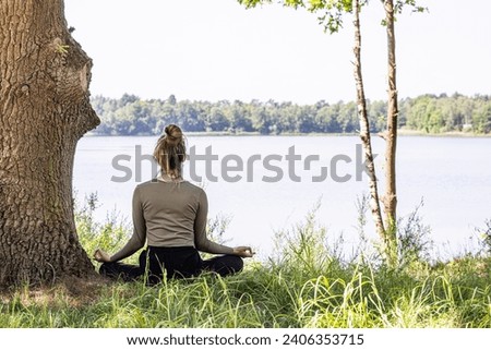A young woman finds inner peace as she practices yoga in the heart of nature. Sitting gracefully on the lush green grass beside a majestic tree and a tranquil forest lake, she engages in a meditation