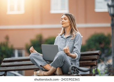 Young woman finds tranquility as she meditates with her laptop on a park bench, blending technology with mindfulness in the urban landscape.