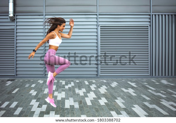 Young woman with fit body jumping and running\
against grey background. Female model in sportswear exercising\
outdoors. Modern young woman in sports clothing jumping while\
exercising outdoors