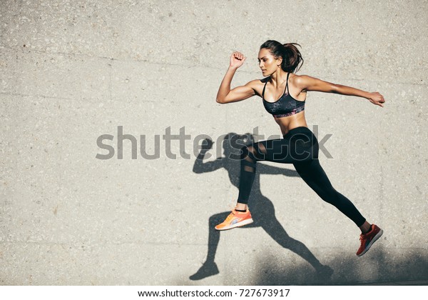 Young\
woman with fit body jumping and running against grey background.\
Female model in sportswear exercising\
outdoors.