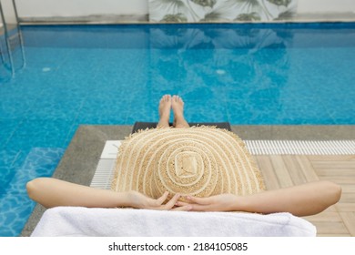 Young Woman With Fit Body Wearing Purple Bikini Swimsuit And Covering Her Face With Broad Brim Straw Hat As Protection From The Sun. Sunbathing By The Swimming Pool. Copy Space.