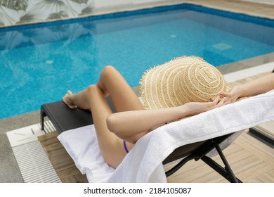 Young Woman With Fit Body Wearing Purple Bikini Swimsuit And Covering Her Face With Broad Brim Straw Hat As Protection From The Sun. Sunbathing By The Swimming Pool. Copy Space.