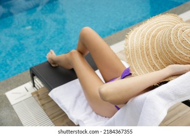 Young Woman With Fit Body Wearing Purple Bikini Swimsuit And Covering Her Face With Broad Brim Straw Hat As Protection From The Sun. Sunbathing By The Swimming Pool. Copy Space.