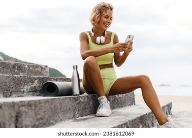 Young woman in fitness clothing sits on seaside stairs, using her phone before her yoga session by the beach, embracing the serene atmosphere and preparing to start her workout routine. - Powered by Shutterstock