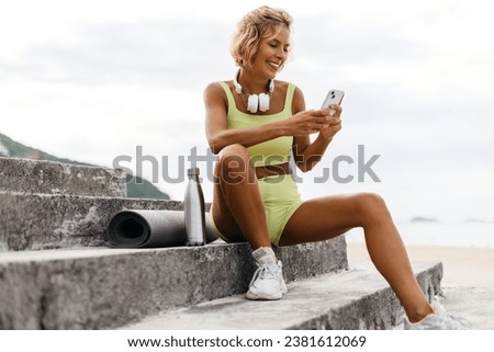 Young woman in fitness clothing sits on seaside stairs, using her phone before her yoga session by the beach, embracing the serene atmosphere and preparing to start her workout routine.