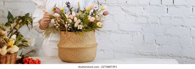 A young woman florist in white clothes and a straw hat stands with a basket of flowers against a white brick wall, in a flower shop. Banner for website header design with copy space.