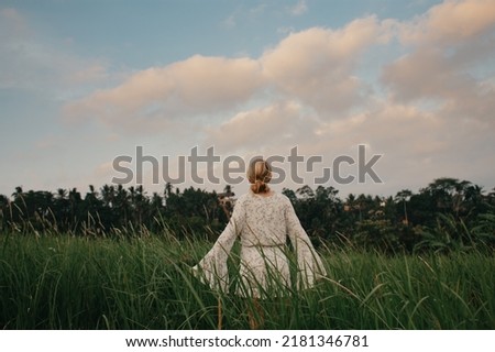 Young woman in flown white dress walking through tall grass field