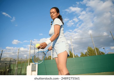 Young woman focusing on serving a tennis ball on an outdoor court. She is wearing sportswear and holding a tennis racket under a partly cloudy sky. - Powered by Shutterstock