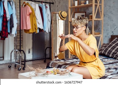 Young woman food blogger wearing eyeglasses sitting at stylish urban apartment taking photo of plate with desserts macaroons and eclairs on smartphone smiling joyful
