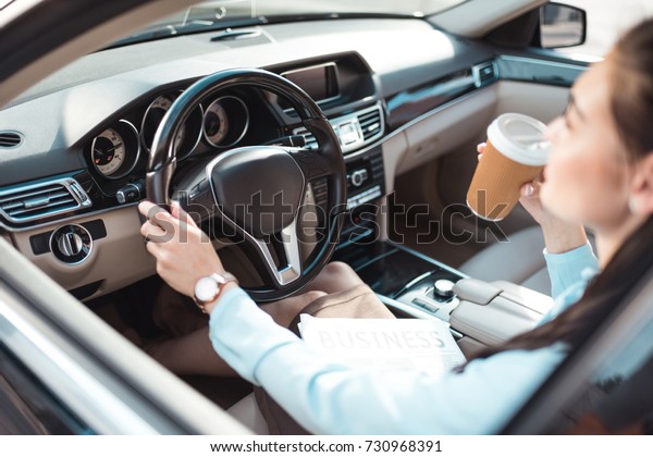 Young woman in formal wear driving a car and\
holding a cup of coffee