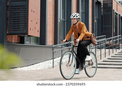 Young woman in formalwear and safety helmet sitting on bicycle and riding home from work while moving along modern building