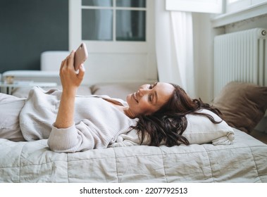 Young Woman Forty Year With Brunette Long Hair In Cozy Knitted Cardigan Using Mobile Phone In Bed At Home