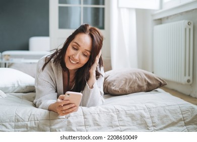 Young Woman Forty Year With Brunette Long Hair In Cozy Knitted Cardigan Using Mobile Phone In Bed At Home