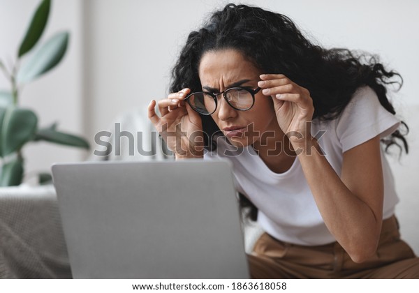 Young woman freelancer with bad eyesight using
laptop, trying to work from home, copy space. Curly lady holding
her glasses and squinting, looking at laptop screen, having vision
troubles